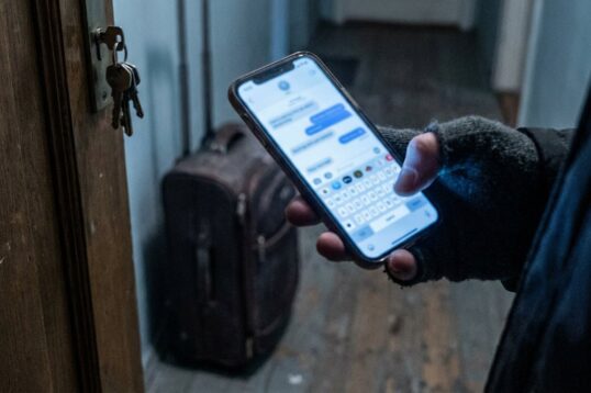 Un voyageur écrit à l’assistance Airbnb depuis son téléphone, devant la porte d’un logement dont les clés sont restées dans la serrure